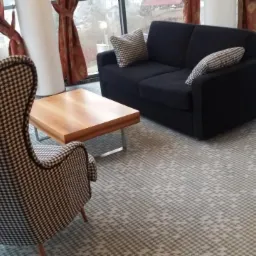 Wnętrze z szarą wykładziną w geometryczny wzór, czarną sofą, fotelem w pepitkę i drewnianym stolikiem kawowym na tle okna.