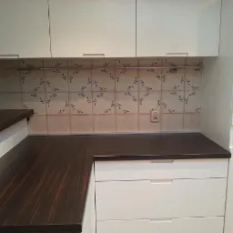 Ciemny drewniany blat kuchenny, kremowe szafki z poziomymi uchwytami i dekoracyjne płytki ceramiczne z motywem roślinnym nad blatem.