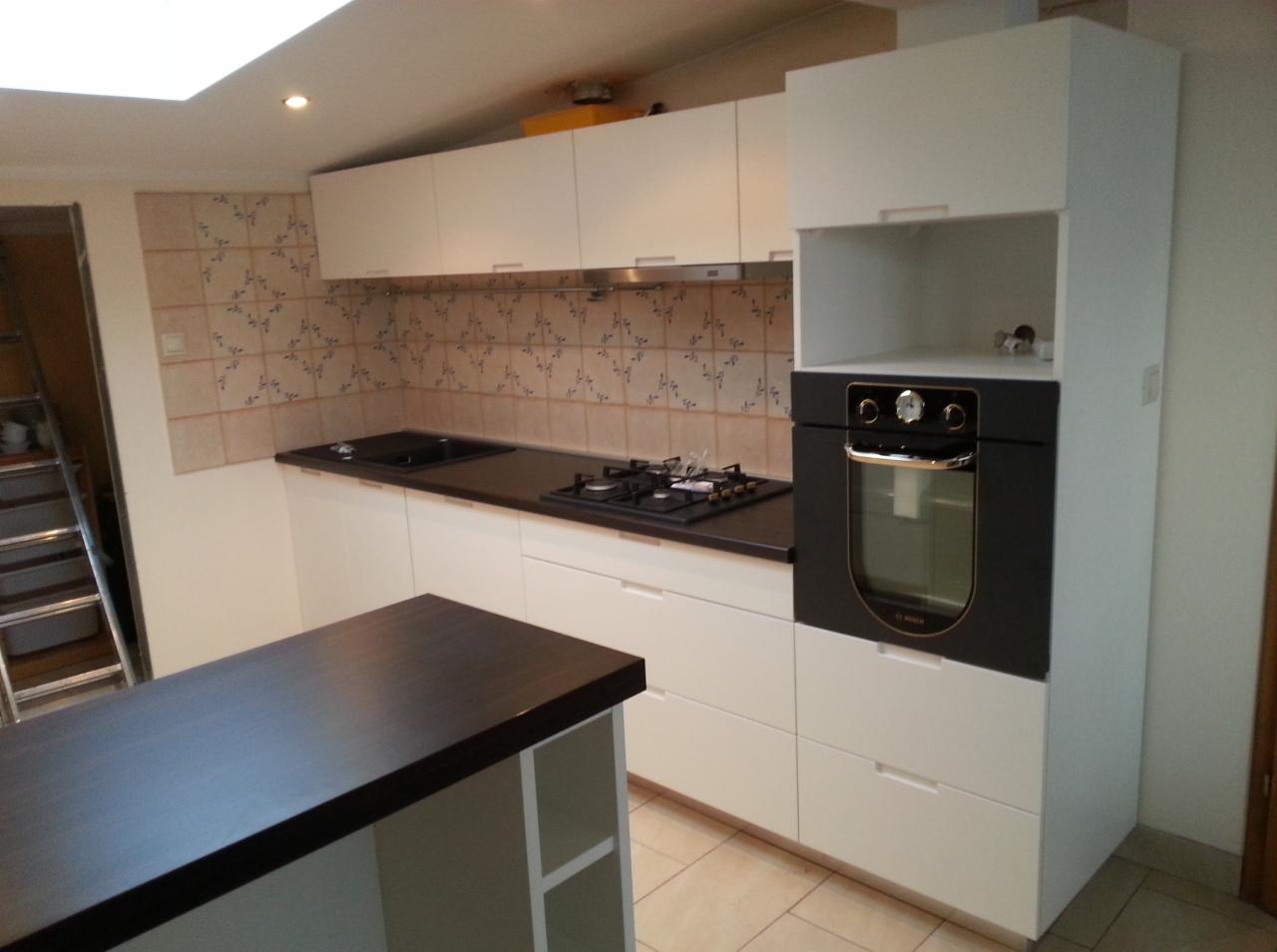 Jasna kuchnia z białymi szafkami i ciemnym blatem, widok na wyspę kuchenną i zabudowany piekarnik. Na ścianie płytki z delikatnym wzorem.