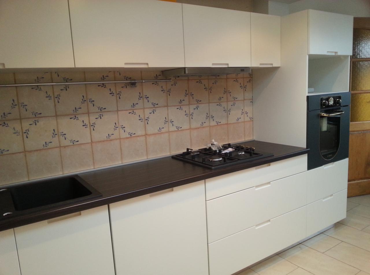 Jasna zabudowa kuchenna z ciemnym blatem i płytą gazową. Nad blatem płytki z motywem roślinnym. Widoczny piekarnik w zabudowie i szafki wiszące.