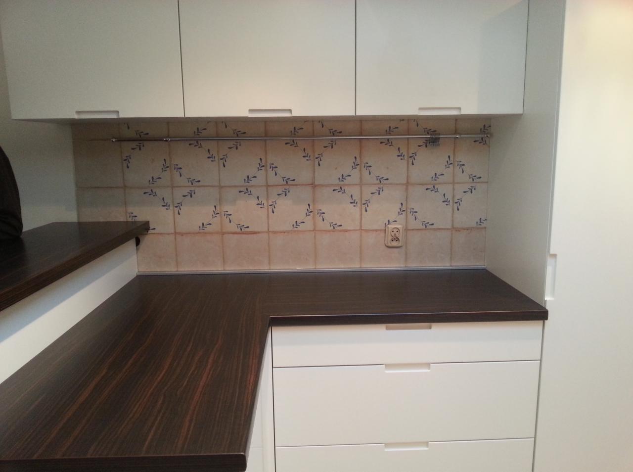 Ciemny drewniany blat kuchenny, kremowe szafki z poziomymi uchwytami i dekoracyjne płytki ceramiczne z motywem roślinnym nad blatem.