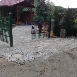 Ułożona z kostki brukowej ścieżka prowadząca do otwartej zielonej bramy, widoczny fragment domu z zadaszeniem i osoba przechodząca przez bramę.