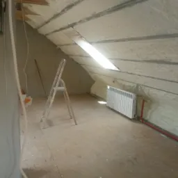 Poddasze po ociepleniu pianką: widoczne białe wypełnienie piankowe na skośnych ścianach i suficie, aluminiowa drabina po lewej stronie, grzejnik na ścianie z pianką, okno dachowe w suficie.