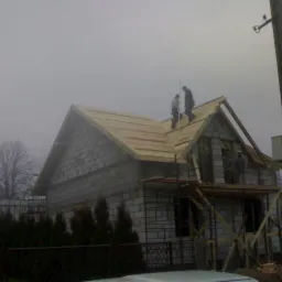 Montaż więźby dachowej w Niegowie na surowym domu z bloczków. Ekipa na dachu, rusztowania. Widok na budynek w trakcie prac.