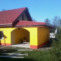 Żółty dom z czerwonym dachem, podcień z łukiem, widoczne deski na trawniku. Budynek po remoncie elewacji i dachu, w słoneczny dzień.
