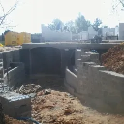 Wykop pod budowę domu z widocznymi fundamentami i ścianami piwnicznymi w trakcie wznoszenia. Na drugim planie materiały budowlane i częściowo wzniesione ściany parteru.