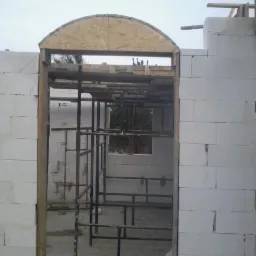 Wznoszenie ściany z bloczków, widoczny fragment konstrukcji łukowej nad otworem. Na drugim planie rusztowanie i fragment ściany z oknem.