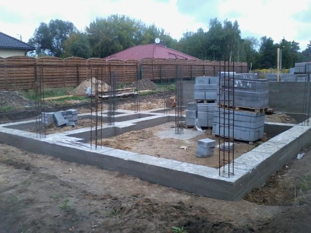 Wylane fundamenty domu z widocznym zbrojeniem i paletami bloczków betonowych. Na dalszym planie ogrodzenie i dom z czerwonym dachem.