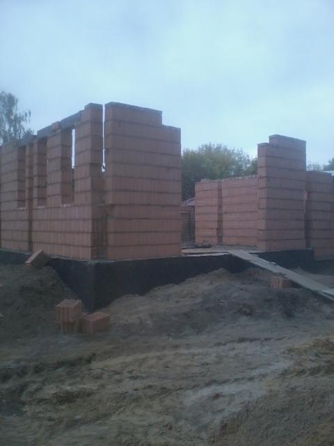 Murowanie ścian budynku z czerwonej cegły w Niegowie. Widoczne fragmenty ścian i fundamenty. Budowa w toku, teren wokół rozkopany.