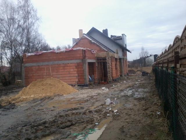 Wznoszenie dwukondygnacyjnego domu z czerwonej cegły. Widoczny fragment dachu i okna. Na pierwszym planie hałda piasku i błotniste podłoże.