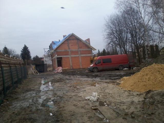 Budowa domu z czerwonej cegły w trakcie realizacji, częściowo przykryta folią. Czerwony bus na posesji, błotniste podłoże. Widok z zewnątrz, pochmurny dzień.