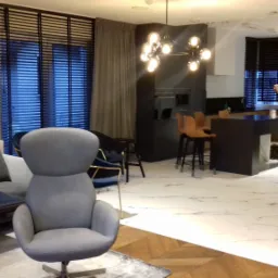 Nowoczesny salon z szarym fotelem, sofą i ciemnymi żaluzjami okiennymi, widoczna kuchnia z wyspą i oświetleniem punktowym, wykończenie pod klucz.