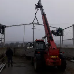 Montaż konstrukcji stalowej dachu hali z użyciem podnośnika teleskopowego Manitou, pracownicy na rusztowaniu i w koszu podnośnika, pochmurny dzień.
