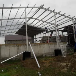 Budowa stalowej konstrukcji dachu w Głubczycach, widoczne elementy więźby dachowej, pracownicy na wysokościach, silos zbożowy w tle, betonowa podmurówka.