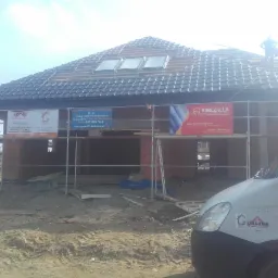 Montaż dachówki ceramicznej na nowym domu w Głubczycach, widoczne okna dachowe i rusztowanie z banerami reklamowymi firm budowlanych.