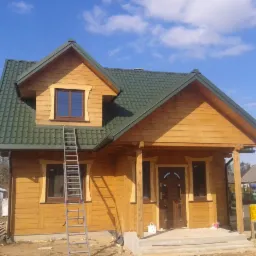 Nowo wybudowany, parterowy dom z jasnego drewna z zielonym dachem, drabina oparta o ścianę, widoczne detale wykończenia i otoczenie w trakcie zagospodarowywania.