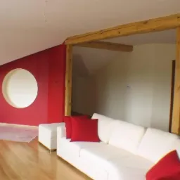 Poddasze po remoncie: czerwona ściana z okrągłym otworem, drewniane belki stropowe, biała sofa z czerwonymi poduszkami, zabezpieczona folią podłoga.