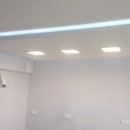 Wykończony sufit podwieszany z płyt kartonowo-gipsowych z wbudowanym oświetleniem LED, widoczne trzy kwadratowe lampy i listwa LED w górnej części. Na ścianie widoczne gniazdka elektryczne i rura...