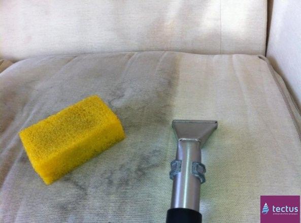 Czyszczenie tapicerki sofy jasnym odkurzaczem piorącym, obok leży żółta gąbka, widoczne ślady po czyszczeniu.