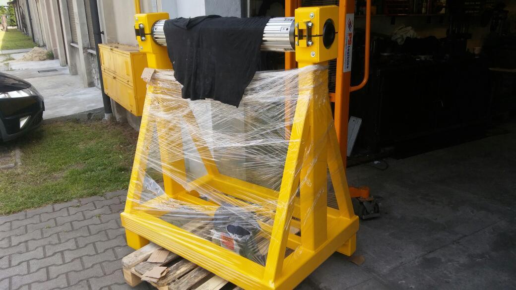 Żółta konstrukcja podtrzymująca metalowy cylinder, owinięta folią stretch, stoi na drewnianej palecie przed budynkiem.