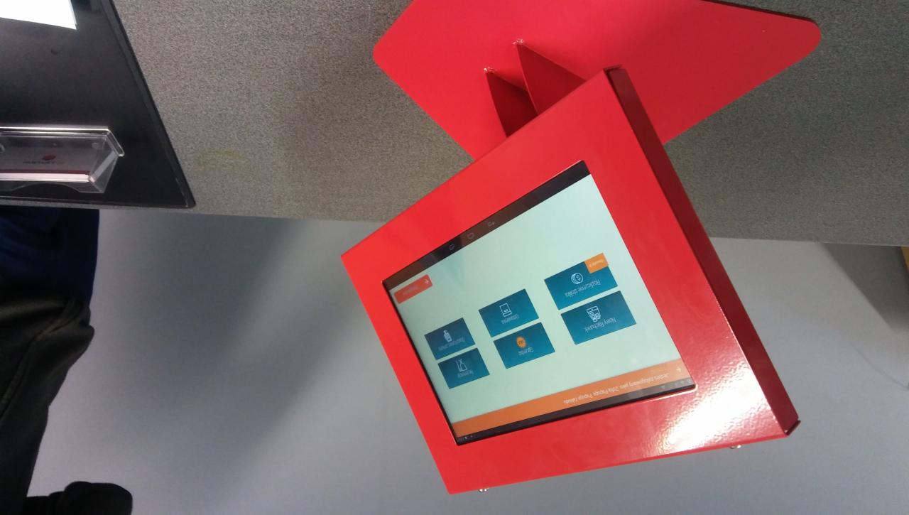 Czerwona, metalowa obudowa interaktywnego kiosku informacyjnego z ekranem dotykowym, prezentującego menu z ikonami i napisami w języku polskim, zamontowana na ścianie.