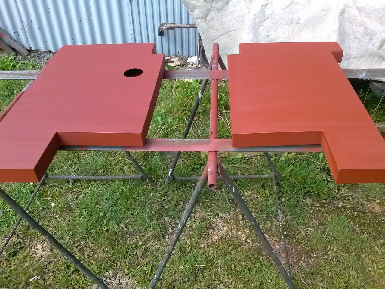 Dwie pomalowane na czerwono, metalowe płyty o nieregularnym kształcie, leżące na metalowym stojaku na trawie. Jedna płyta ma okrągły otwór.