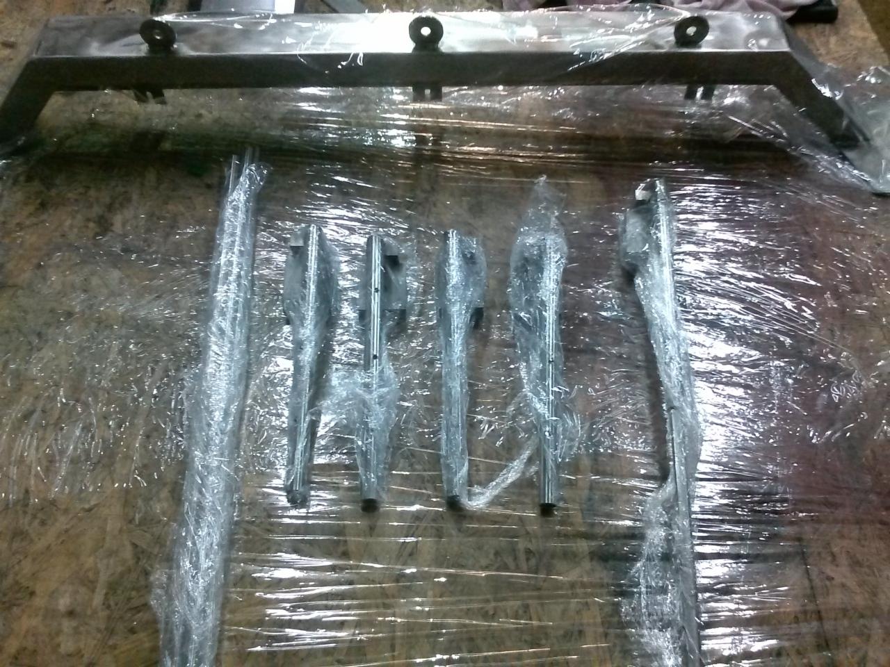 Metalowe elementy konstrukcyjne, w tym rama z uchwytami i pręty, owinięte folią ochronną na drewnianej powierzchni.