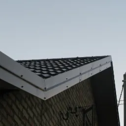 Ciemna dachówka ceramiczna na skośnym dachu budynku z cegły, widok z dołu, fragment rynny i słup elektryczny w tle.