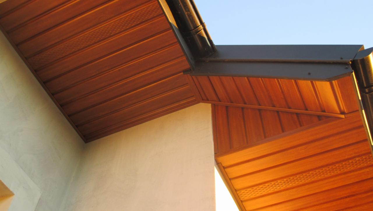 Detale okapu dachowego z drewnianą podbitką w ciepłym odcieniu brązu i czarną rynną. Widok z dołu, z fragmentem ściany w tle.