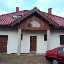 Nowy dom jednorodzinny z czerwoną dachówką i garażem, widoczny częściowo czerwony samochód. Świeżo otynkowana elewacja, okna i drzwi w kolorze brązowym.