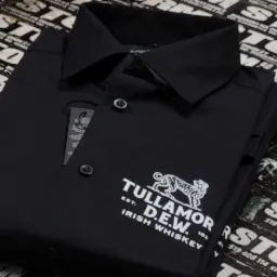 Złożona czarna koszula polo z białym nadrukiem logo Tullamore D.E.W. Irish Whiskey na tle ulotek reklamowych firmy STORM.