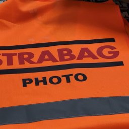 STORM STREETWEAR ŁUKASZ JAWORSKI - Pomarańczowa kamizelka odblaskowa z czarnym nadrukiem 'STRABAG PHOTO' na tle zadrukowanych materiałów reklamowych.