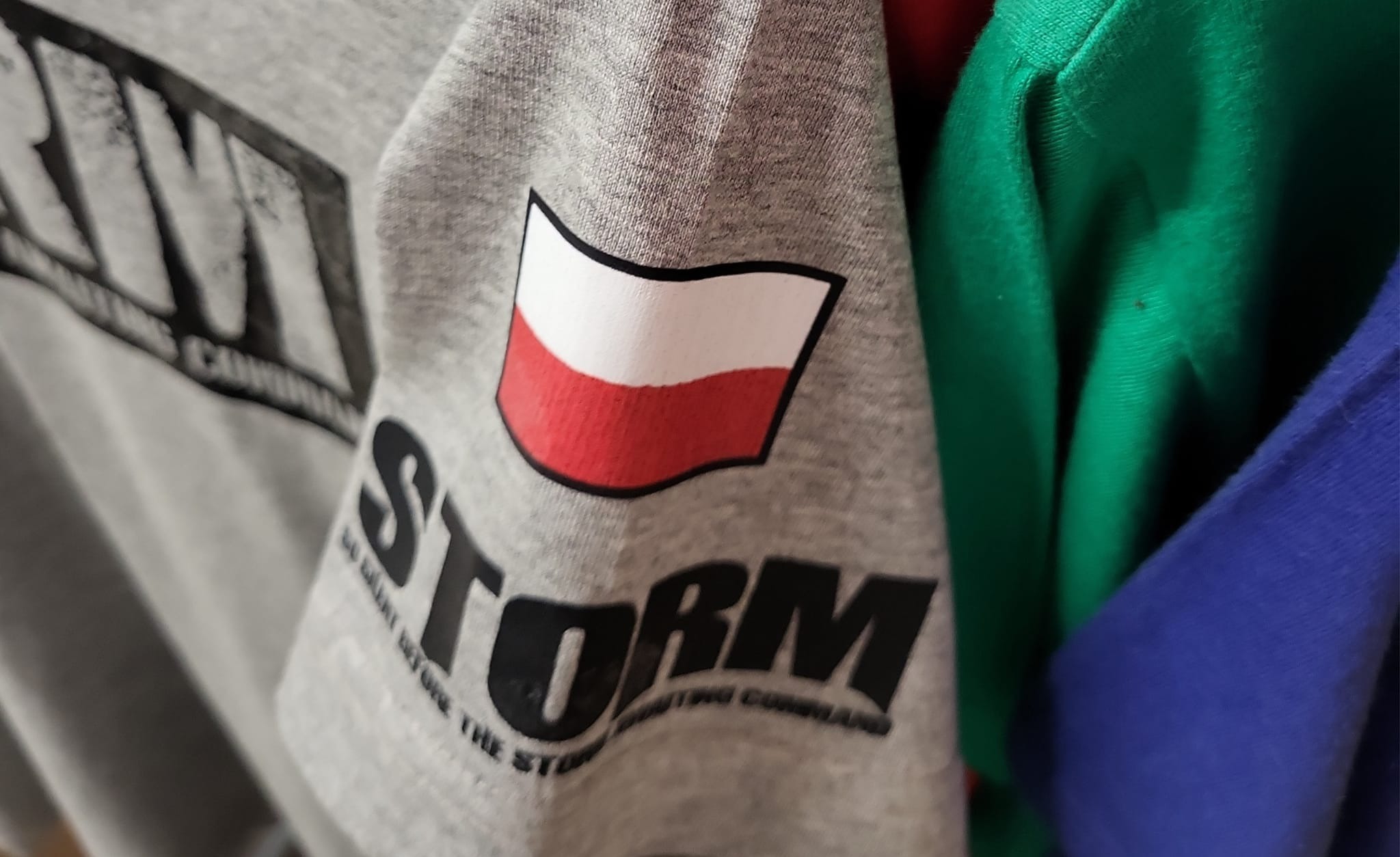 Szara koszulka z nadrukiem flagi Polski i napisem STORM na tle innych kolorowych ubrań.