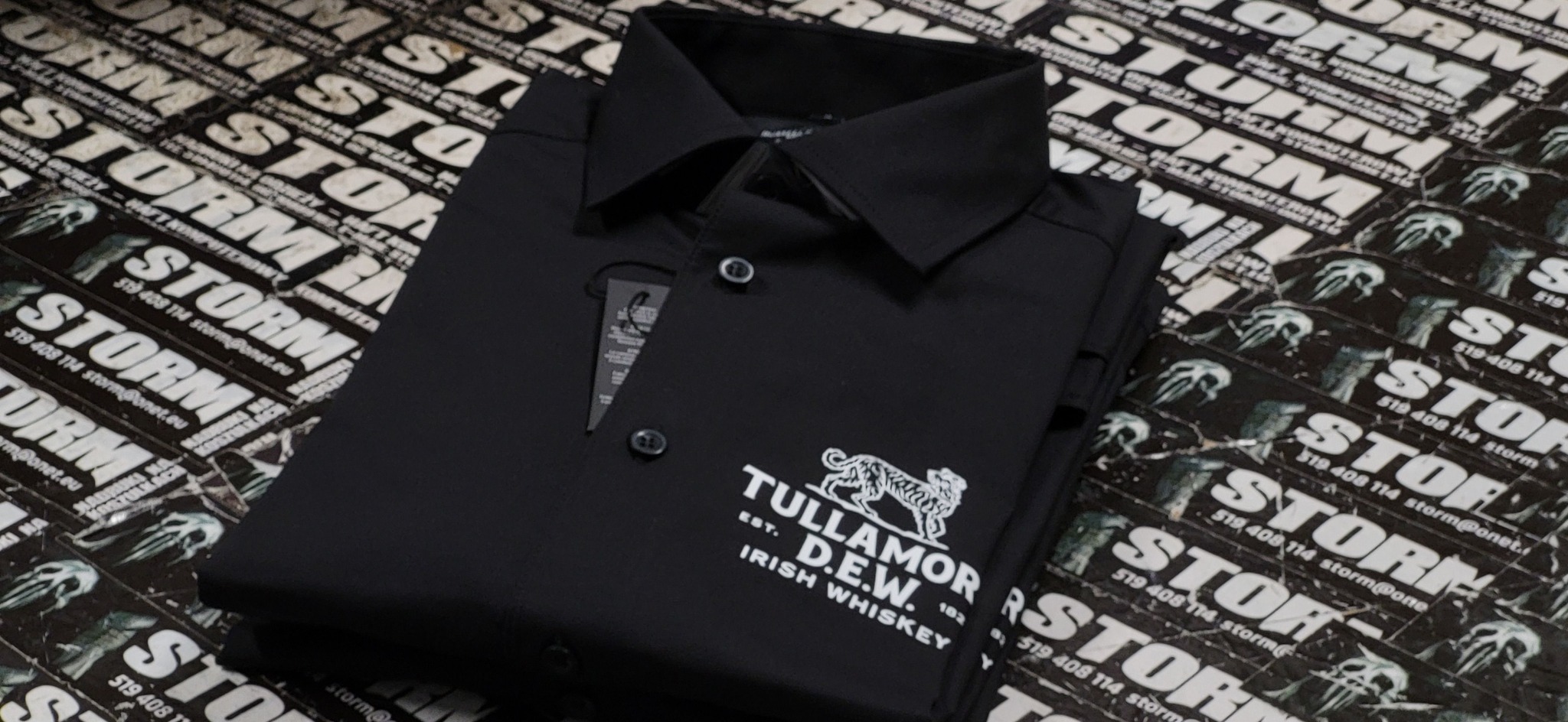 Złożona czarna koszula polo z białym nadrukiem logo Tullamore D.E.W. Irish Whiskey na tle ulotek reklamowych firmy STORM.