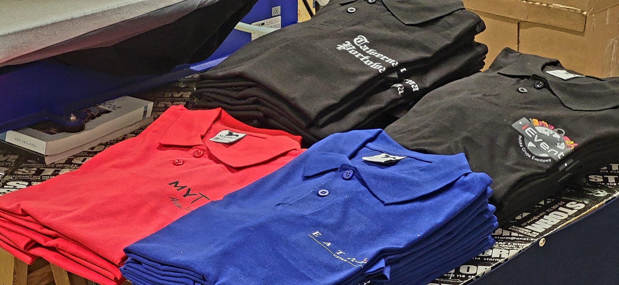Stosy koszulek polo w kolorach czerwonym, niebieskim i czarnym, z widocznymi nadrukami różnych firm i logo, ułożone na stole z reklamami.
