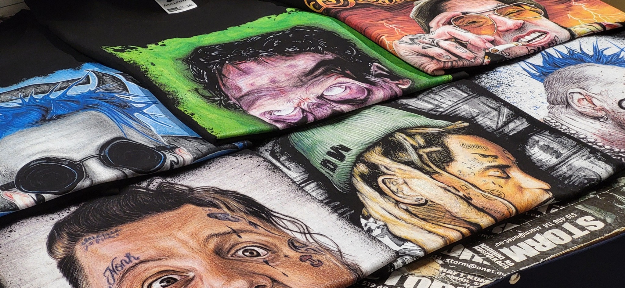 Stos koszulek z nadrukami portretów w różnej stylistyce, od realistycznych po surrealistyczne, ułożonych warstwami, z widocznymi detalami tatuaży i ekspresji twarzy.