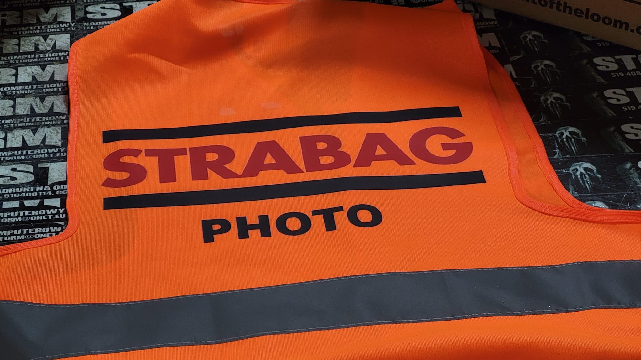 Pomarańczowa kamizelka odblaskowa z czarnym nadrukiem 'STRABAG PHOTO' na tle zadrukowanych materiałów reklamowych.