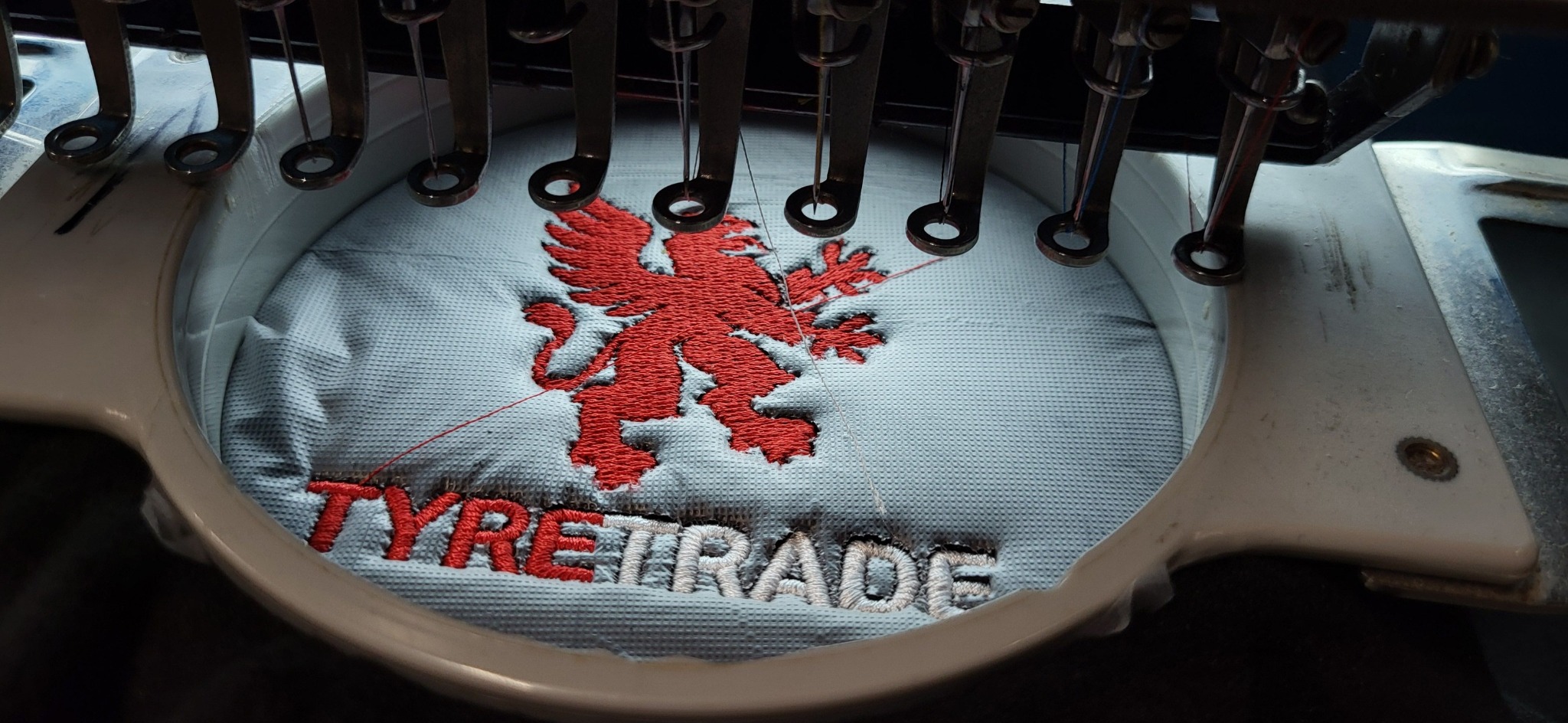 Proces haftowania czerwonego lwa i napisu TYRE TRADE na jasnoniebieskim materiale za pomocą maszyny do haftowania.