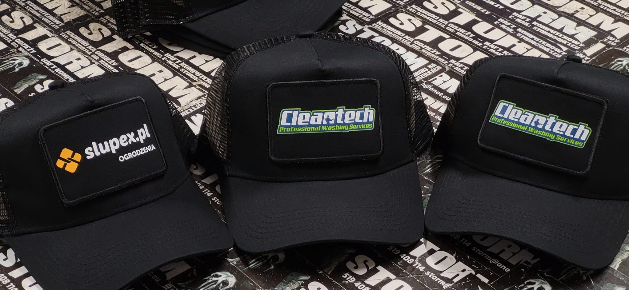 Trzy czarne czapki typu trucker z siatką, każda z naszywką z logo innej firmy: slupex.pl z branży ogrodzeń oraz Cleartech Professional Washing Services, leżące na tle z nadrukami reklamowymi.