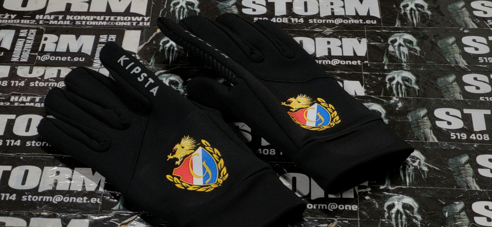 Para czarnych rękawic sportowych marki Kipsta z wyhaftowanym herbem, leżących na tle plakatów z logo firmy Storm.