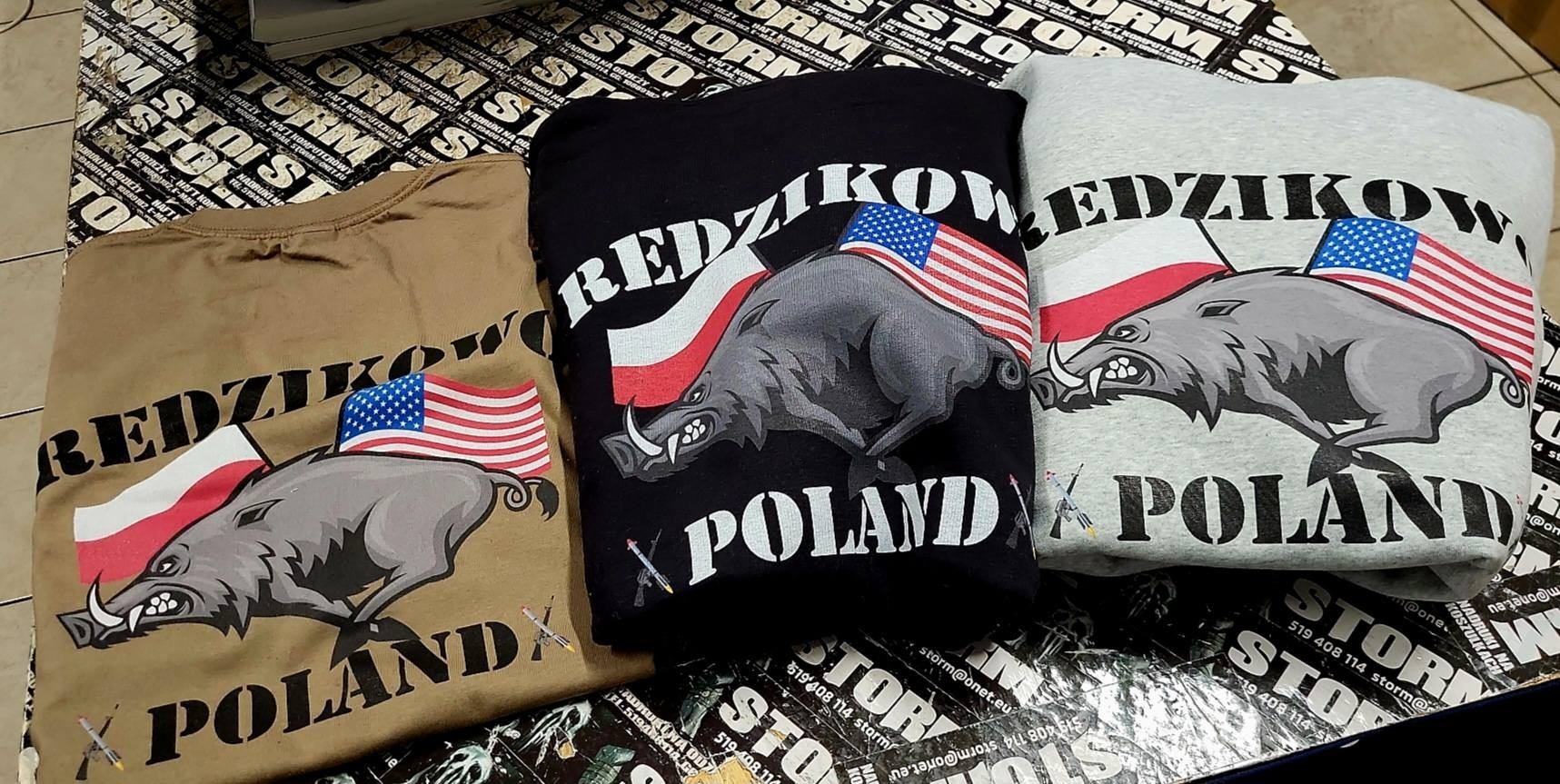 Trzy koszulki w kolorach khaki, czarnym i szarym, z nadrukiem wizerunku dzika, flagi polskiej i amerykańskiej oraz napisem 'REDZIKOW' i 'POLAND', leżące na tle z ulotkami.