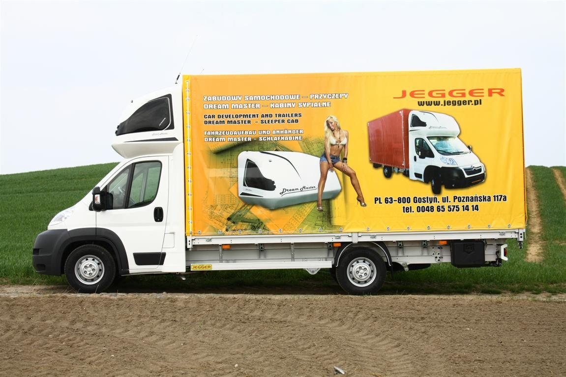 Biały samochód dostawczy z żółtą zabudową skrzyniową reklamującą kabiny sypialne Dream Master, z wizerunkiem kobiety w bikini, na tle zielonego pola.