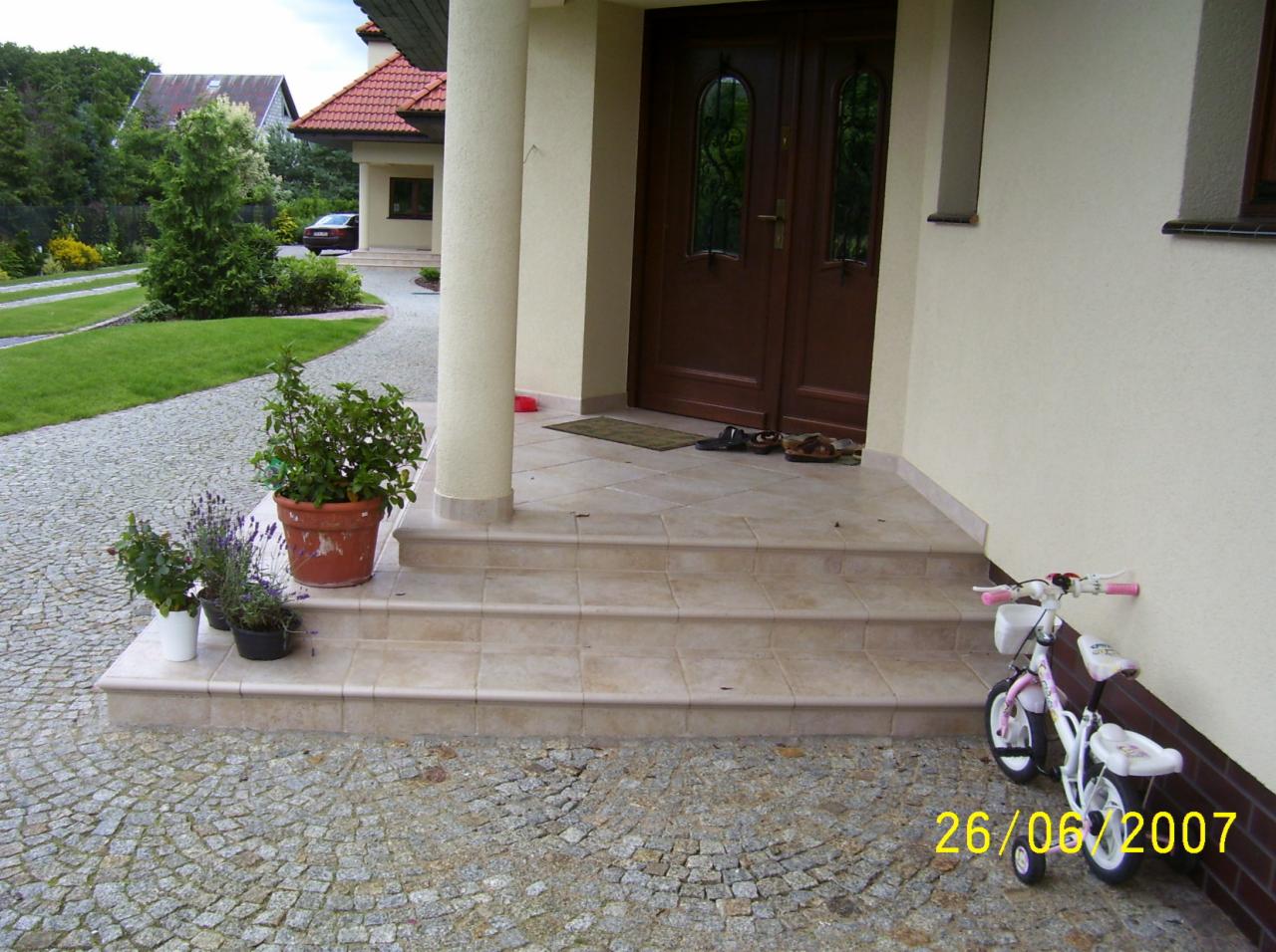 Wyłożone jasnymi płytkami schody wejściowe do domu w Szczecinie, z kolumną i ciemnymi drzwiami, z doniczkami z kwiatami po lewej i dziecięcym rowerkiem po prawej stronie.