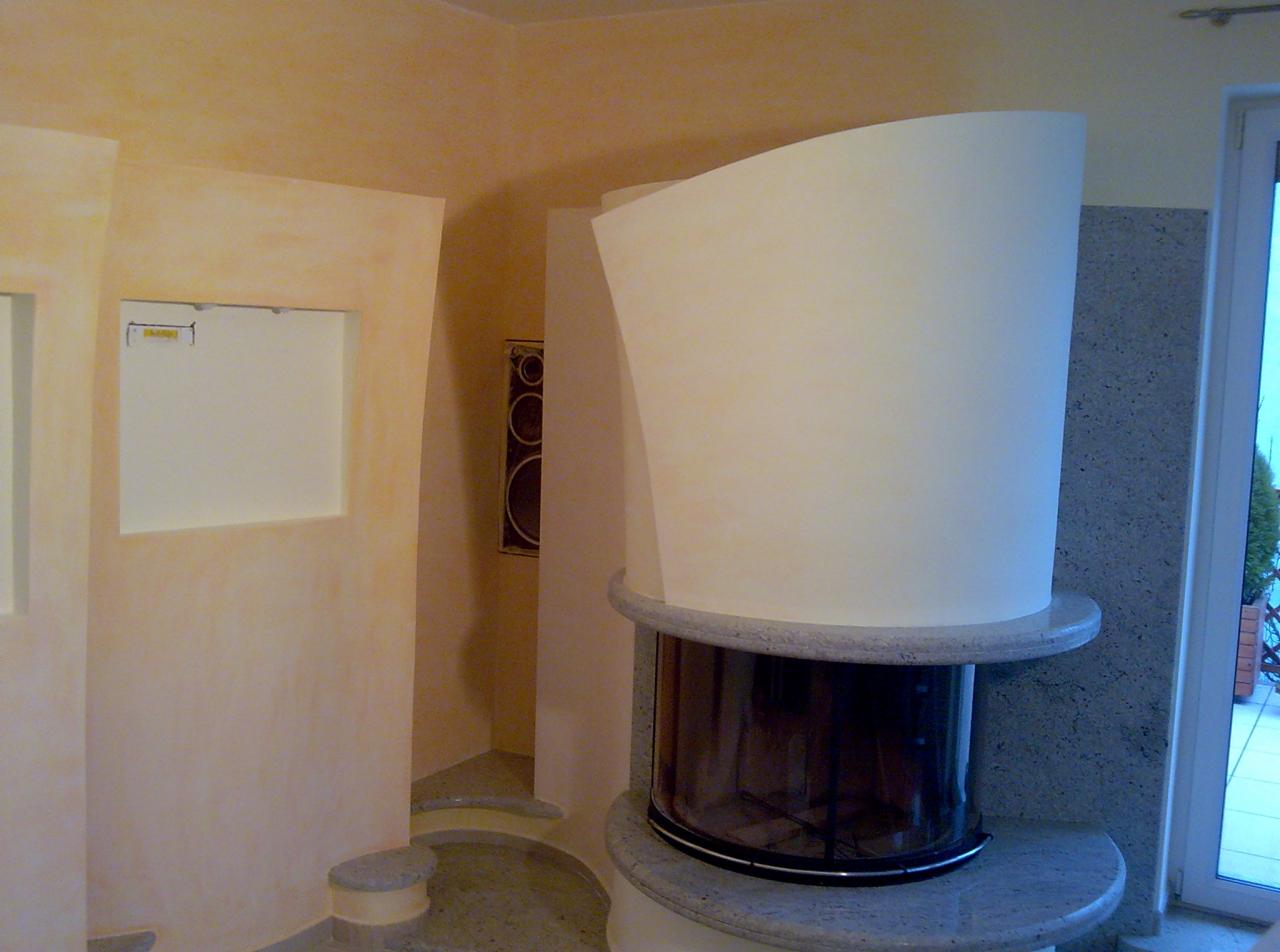 Nowoczesny kominek z obudową w kształcie walca, wykończony granitem, w jasnym wnętrzu z beżowymi ścianami i schodkowym przejściem.