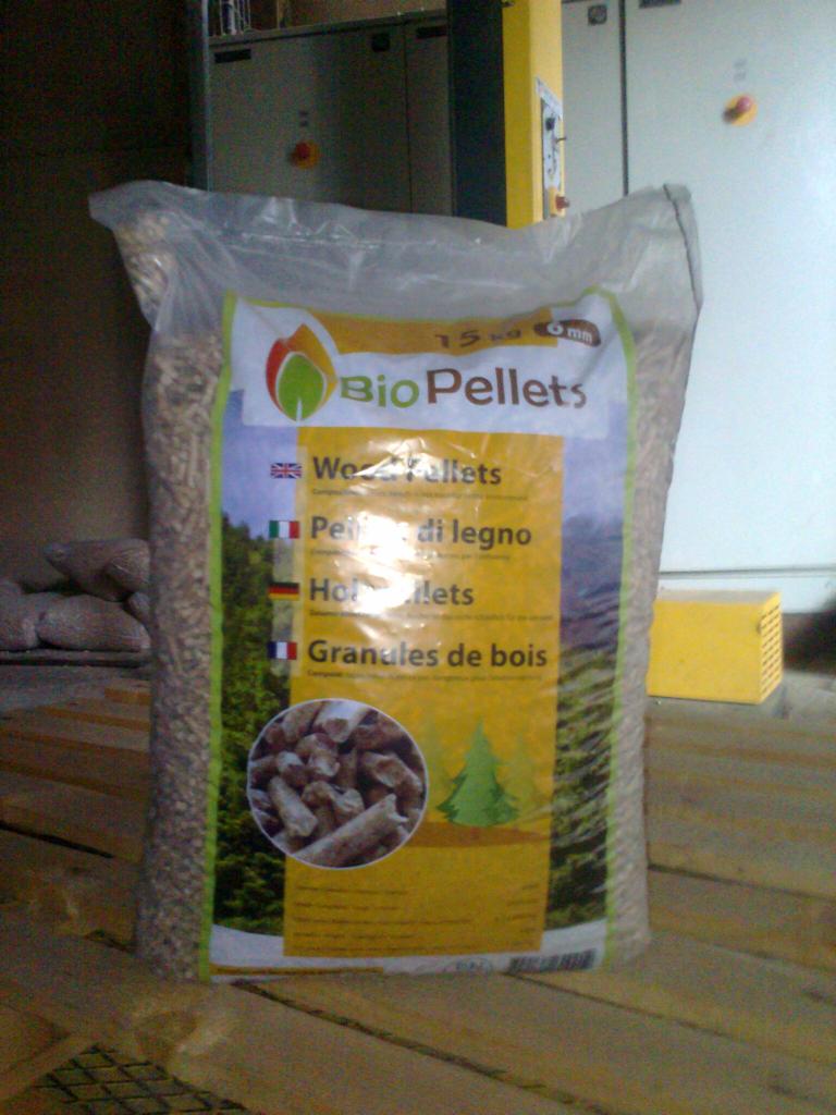 Worek BioPellets 15 kg, widoczne tłumaczenia nazwy na różne języki, na drewnianej podłodze, w tle szafa sterownicza.