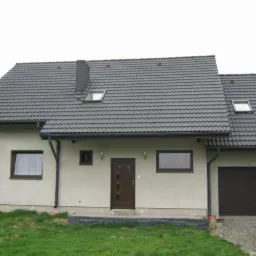 Dom jednorodzinny z grafitowym dachem i brązowymi oknami. Widoczny fragment ogrodu i samochód. Dach z oknami połaciowymi i kominem. Elewacja w odcieniach beżu.