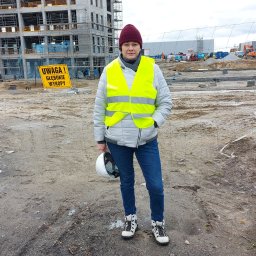 Agata Waszkiewicz Professional - Kobieta w kamizelce odblaskowej i kasku na budowie, w tle konstrukcja budynku i tablica ostrzegawcza o głębokich wykopach. Ujęcie z perspektywy osoby stojącej za ogrodzeniem.