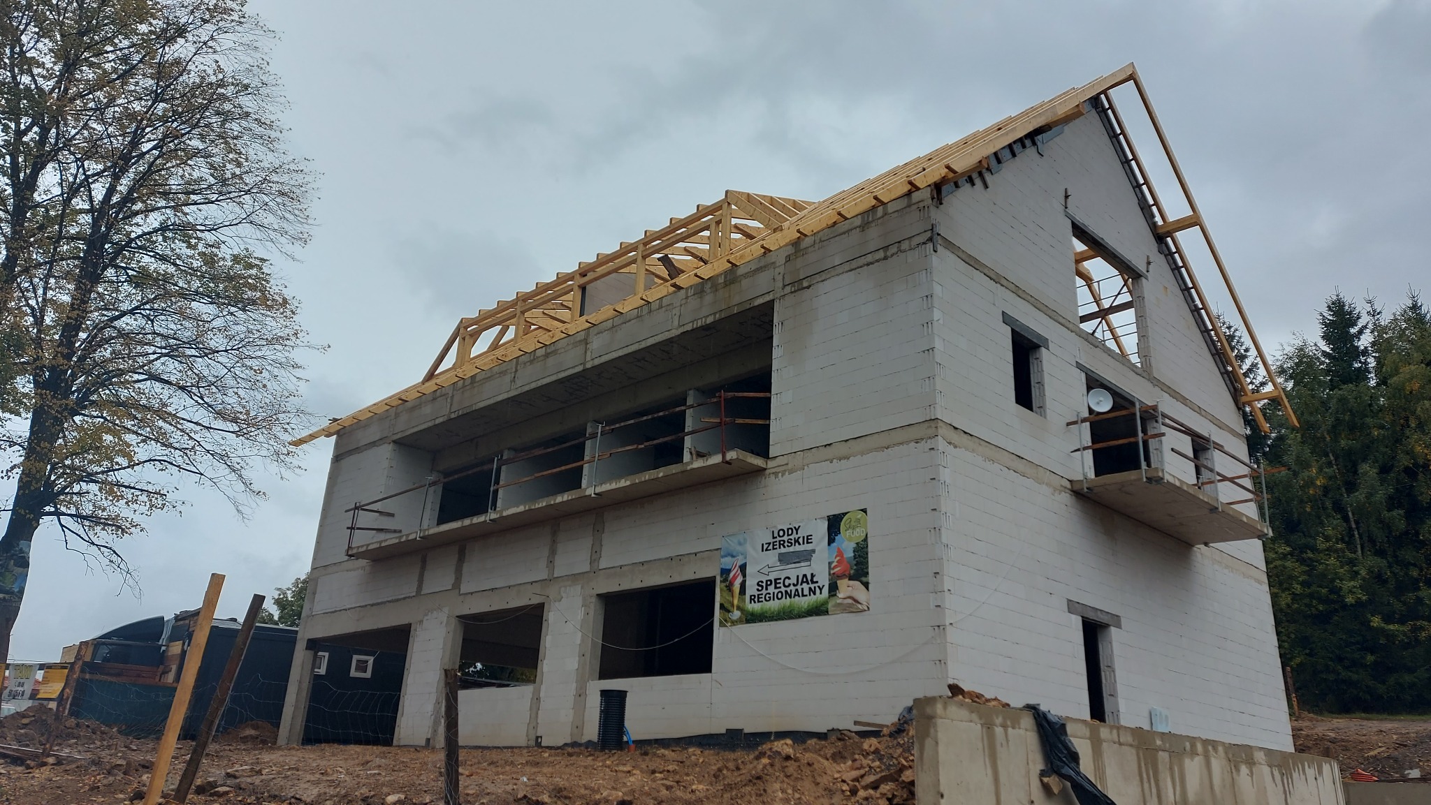 Surowy, dwukondygnacyjny budynek z białego bloczka w trakcie budowy, z widoczną drewnianą konstrukcją dachu i balkonami. Na ścianie baner reklamowy.