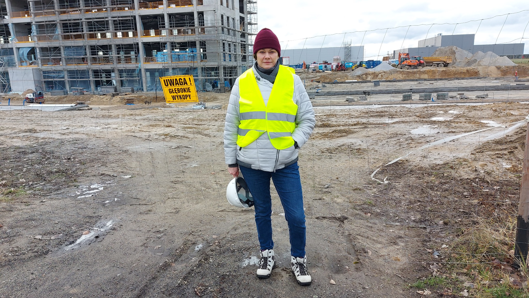 Kobieta w kamizelce odblaskowej i kasku na budowie, w tle konstrukcja budynku i tablica ostrzegawcza o głębokich wykopach. Ujęcie z perspektywy osoby stojącej za ogrodzeniem.