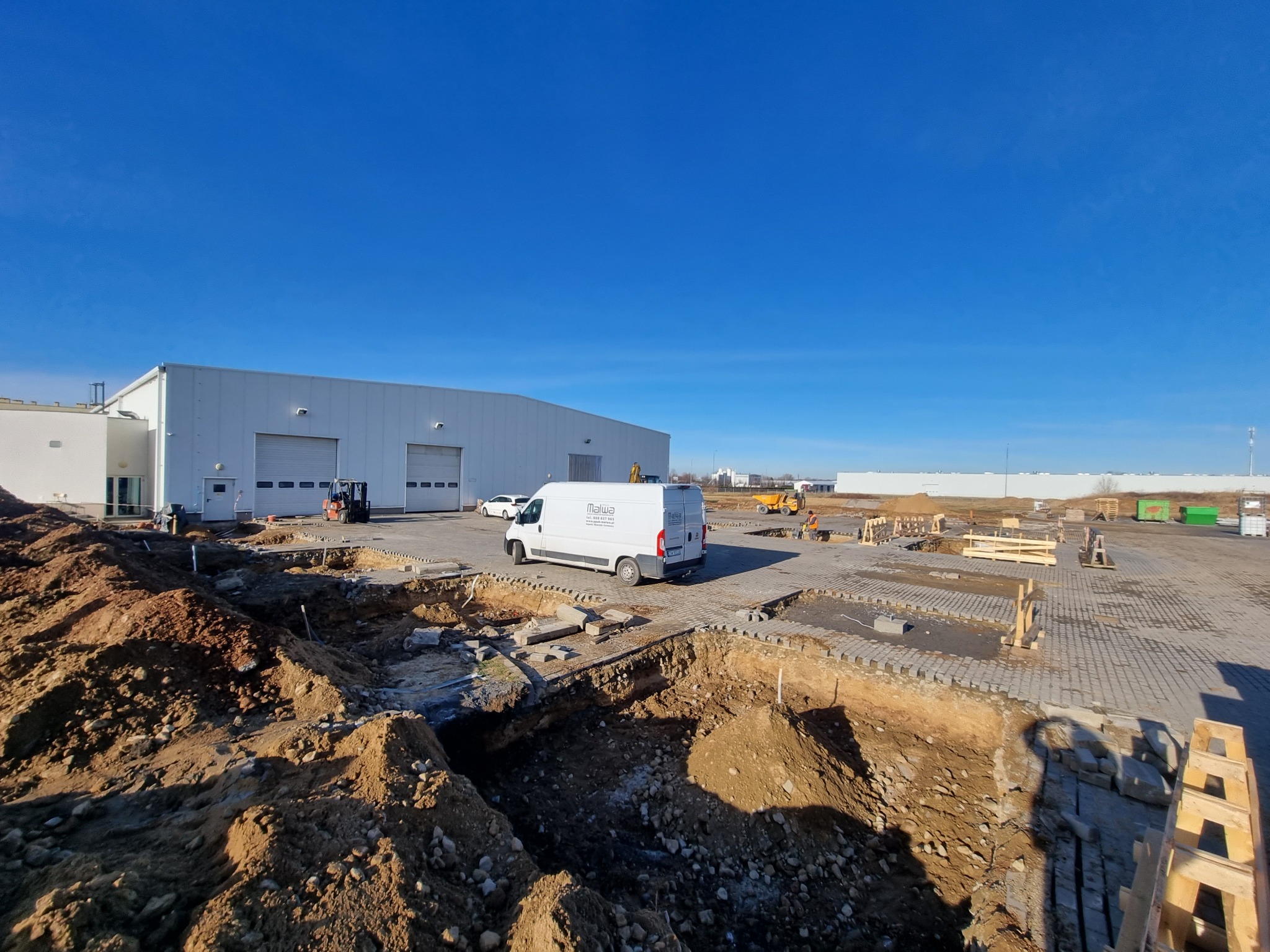 Budowa hali: wykop fundamentów, widoczny biały budynek w tle, firmowy bus i sprzęt budowlany. Ujęcie z góry, podkreślające etap prac ziemnych.